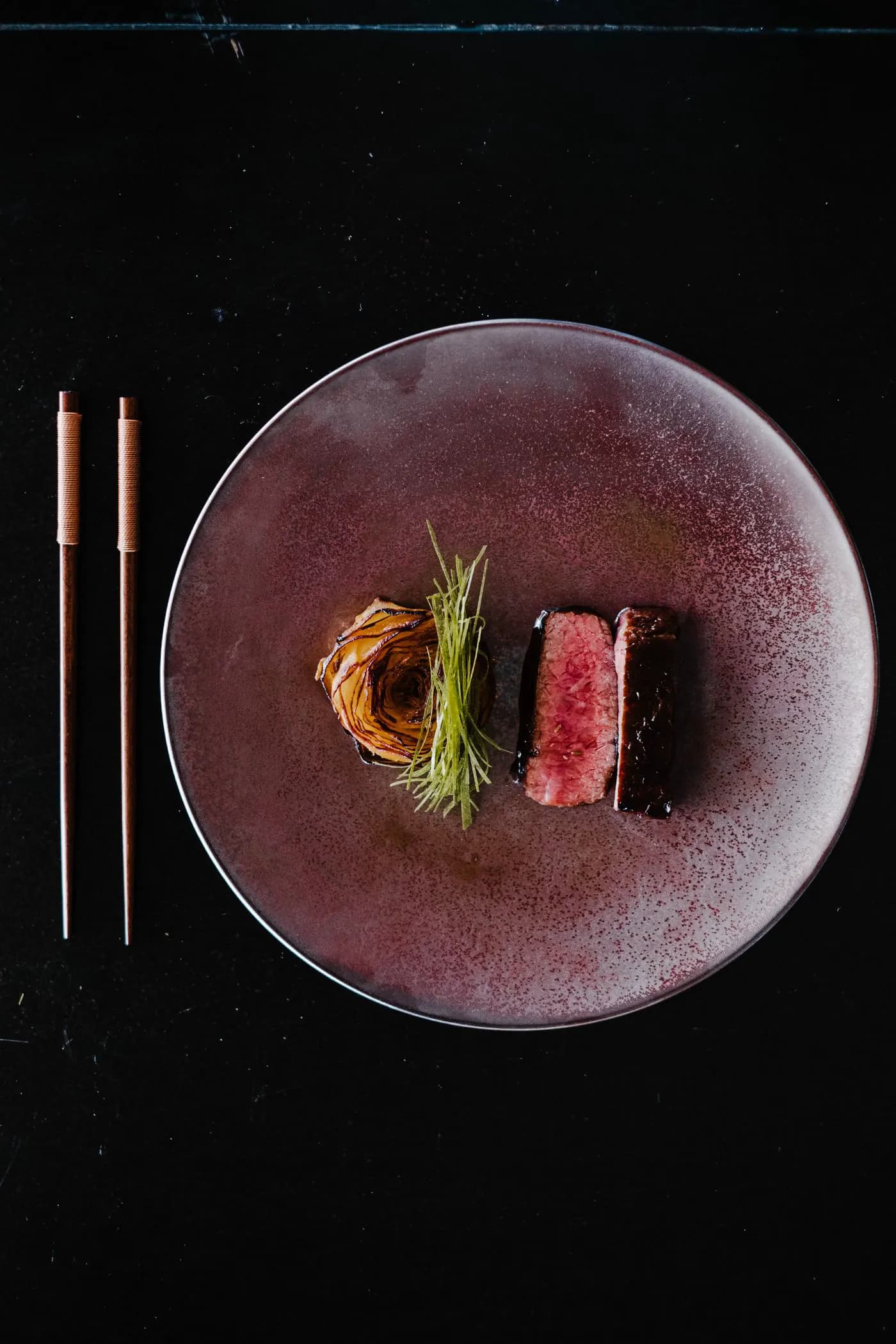 A seared steak and garnish on a dark speckled plate.