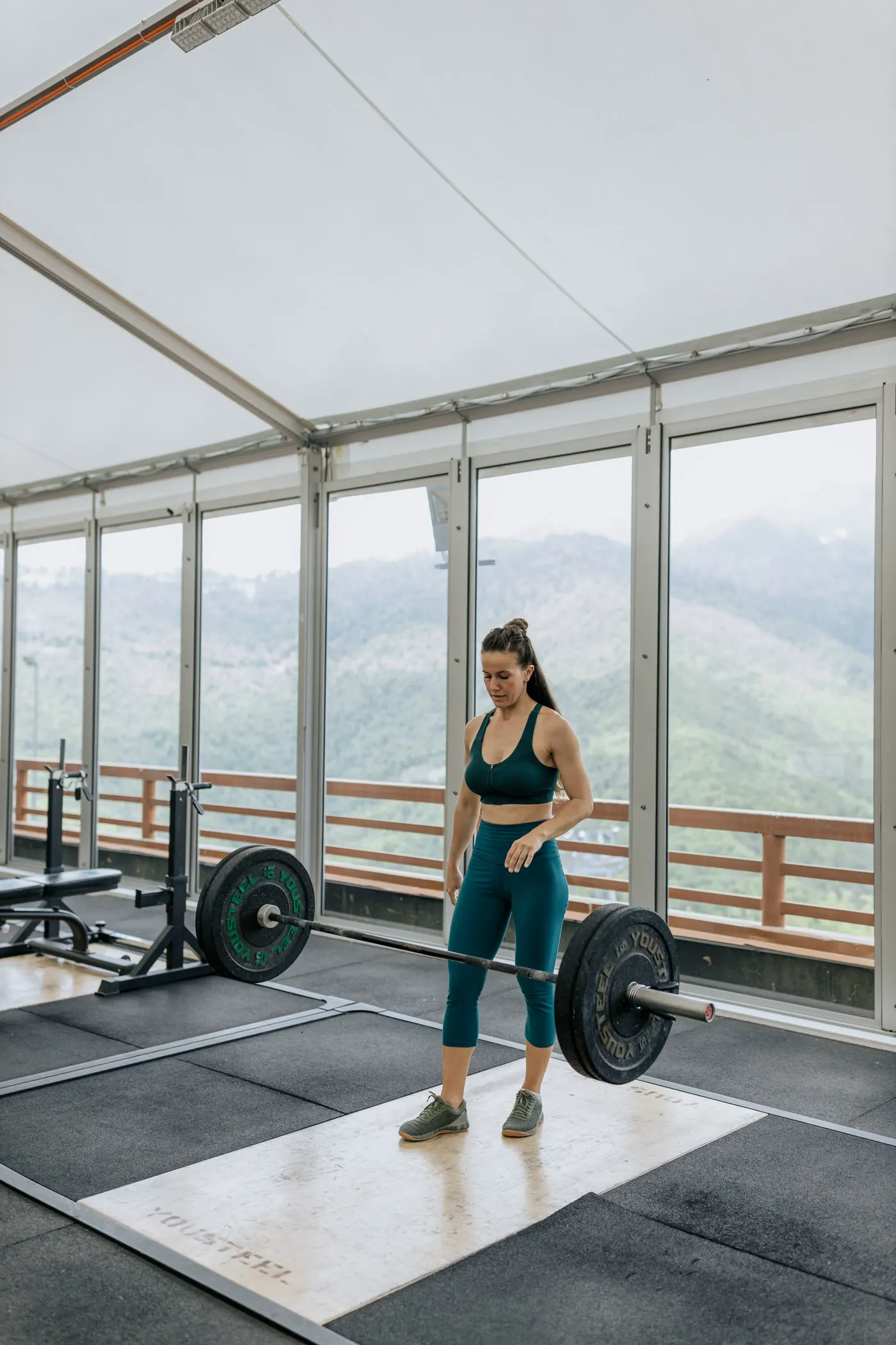 A woman stands ready at a loaded barbell in a bright gym with mountain views.